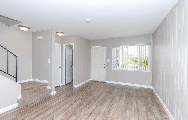 A spacious, empty living room featuring light gray walls, wood-style flooring, and large windows. There's a staircase on the left leading to another level, and a door on the right leading outside. The room has a bright, airy feel with natural light illuminating the space.
