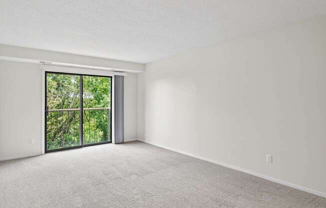 A large empty room with a carpeted floor and a sliding glass door leading to a lush green outdoor area.