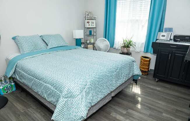 A bed with a blue and white patterned comforter is in the center of a room with a printer and a fan on a desk.