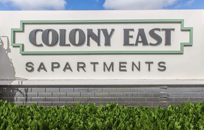 Signage for Colony East Apartments, featuring the name "COLONY EAST" prominently displayed in large letters with decorative elements, situated above the word "APARTMENTS." The sign is positioned against a backdrop of green foliage and a clear blue sky.