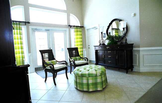 A room with a green and white checkered ottoman, two chairs, a dresser, and a mirror.