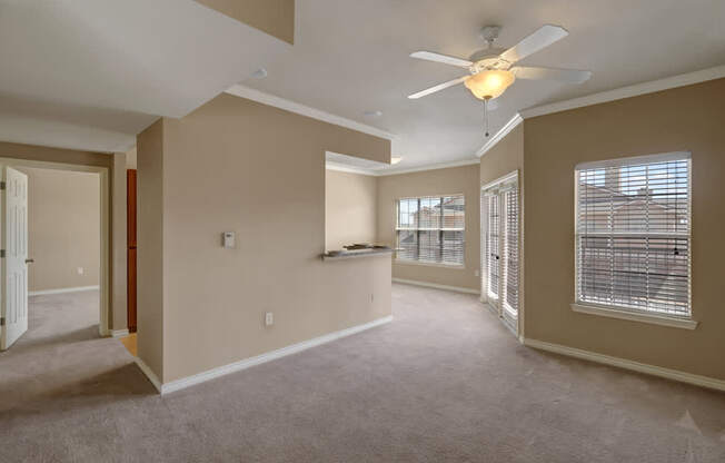 A bright and open living room here at Mission Green featuring plush carpet flooring, neutral wall tones, a ceiling fan with overhead lighting, and multiple windows that bring in natural light, with a pass-through counter to the kitchen and an open layout that connects comfortably to adjacent rooms.