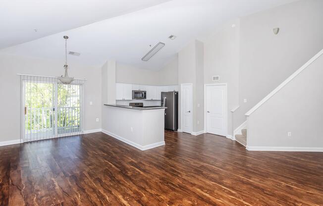 Spacious living area featuring hardwood floors, large windows with natural light, and a breakfast bar connecting to a modern kitchen with stainless steel appliances. The design includes a staircase leading to an upper level, and a sliding glass door opens to a small balcony.