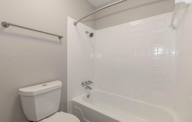 A clean and modern bathroom featuring a white tiled bathtub with a shower rod and curtain, a toilet, and light gray walls. The space is well-lit and has a simple, functional design, making it suitable for everyday use.