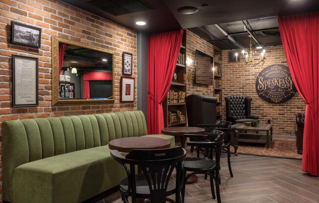 Cozy interior of a speakeasy-style lounge featuring exposed brick walls, a green velvet sofa, wooden tables, and black chairs. A large mirror reflects the ambience, with red curtains and dim lighting adding to the relaxed atmosphere. A sign on the wall reads "Speakeasy," contributing to the vintage aesthetic.