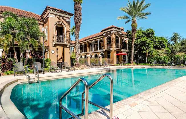 Sparkling swimming pool framed by palm trees and Mediterranean-style buildings.