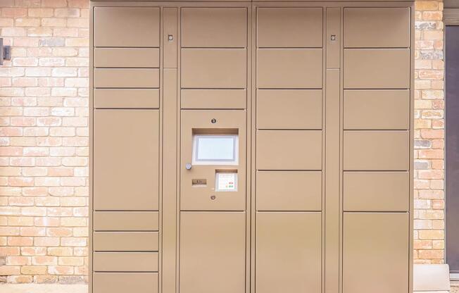 A tan delivery locker unit against a brick wall, featuring a digital screen and buttons for package access. The locker is designed for secure package retrieval, with multiple compartments for different sizes of deliveries.