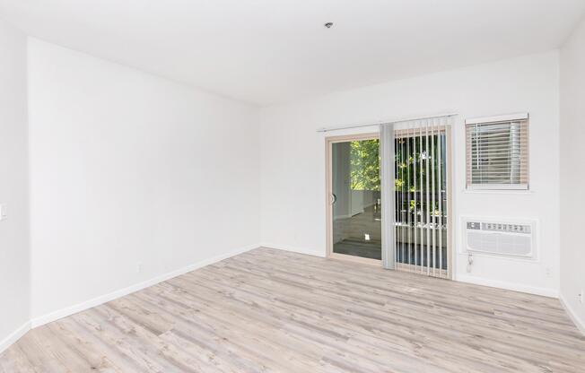 A spacious, empty room with light-colored walls and a wood-like floor. There is a sliding glass door leading to a balcony or patio, accompanied by vertical blinds. An air conditioning unit is mounted on the wall near the door. The room is well-lit by natural light coming through the door.