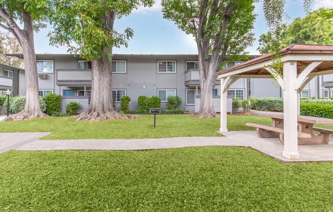 A landscaped area featuring a well-maintained lawn with two large trees, a gazebo with a picnic table, and a pathway leading to apartment buildings in the background. The setting is bright and inviting, with greenery enhancing the peaceful atmosphere.