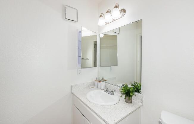 A clean, well-lit bathroom with a white countertop and sink. There is a large mirror above the sink, complemented by three light fixtures. A small potted plant adds a touch of greenery. The walls are painted white, creating a bright and airy atmosphere.