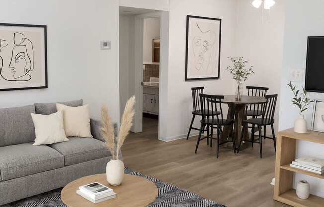 A living room with a grey sofa, a coffee table, a dining table with chairs, and a TV on the wall.