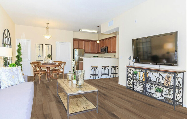 A modern living room with an open kitchen. Features a light-colored sofa with decorative pillows, a wooden coffee table with plants, and a television on a stylish stand. The kitchen has wooden cabinets and bar stools. Natural light fills the space through windows, creating a welcoming atmosphere.