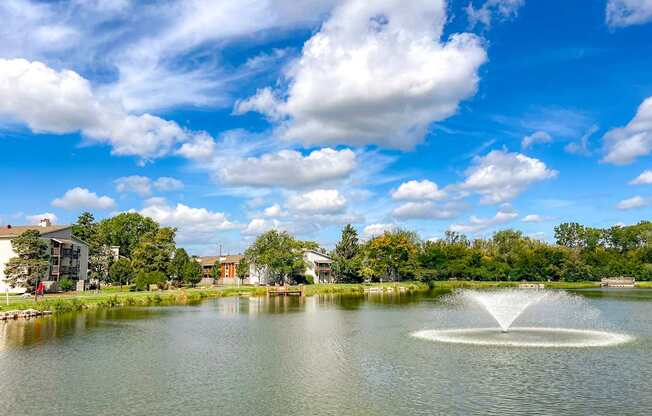 A fountain in the middle of a lake with buildings and trees in the background.