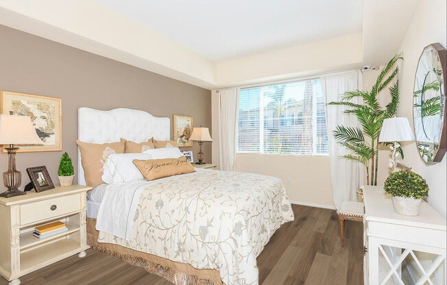 A cozy, well-decorated bedroom featuring a white tufted bed with decorative pillows, a beige and gold quilt, two bedside lamps on stylish nightstands, a window with sheer curtains letting in natural light, and a decorative mirror. A potted plant adds a touch of greenery to the inviting space.