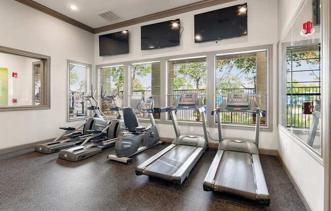 Fitness center with treadmills, bikes, ellipticals, tall windows, and wall-mounted televisions.