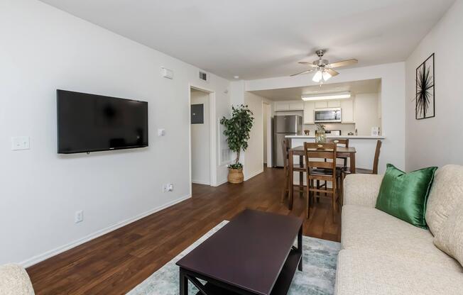 A cozy living room featuring a light-colored sofa with green cushions, a dark wood coffee table, and a flat-screen TV mounted on the wall. Beyond, there's a dining area with a wooden table and chairs, leading to a well-lit kitchen in the background. The space has warm wood flooring and soft lighting.