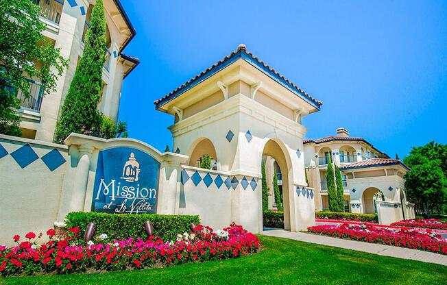 Community entrance with florals and luscious grass at Mission at La Villita Apartments in Irving, TX offers 1, 2 & 3 bedroom apartment homes with appliances.