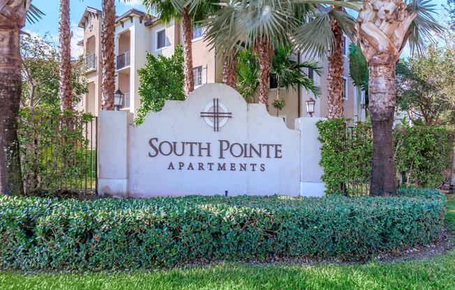 A sign displaying "South Pointe Apartments" surrounded by lush greenery and palm trees, in front of a modern apartment building. The sign is white with decorative elements, set against a backdrop of manicured landscaping.