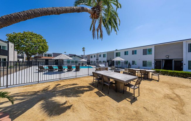 A sandy area with a table and chairs and a pool in the background.