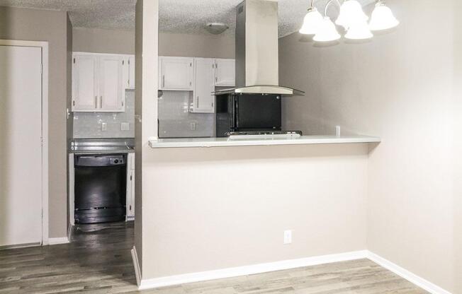 A modern kitchen featuring a bar countertop, black appliances including a dishwasher and stove, and white cabinetry. The space has a light, neutral color palette with wood-style flooring and overhead light fixtures. The open layout connects to the living area.