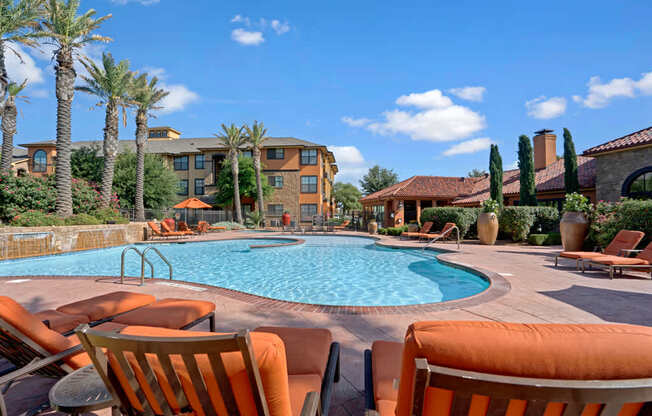 A sunny resort-style swimming pool here at Tuscany at Faudree showcasing a curved pool with clear blue water, surrounding tan pool deck, and orange cushioned lounge chairs in the foreground. Palm trees, landscaped planters, and Mediterranean-inspired buildings frame the pool area beneath a bright blue sky with scattered clouds.