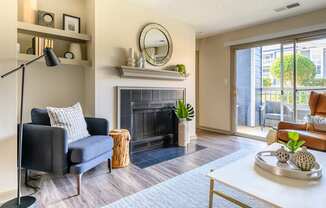 A cozy living room here at Retreat at McAlpine Creek featuring wood-style flooring, a tiled fireplace with a decorative mantel and round mirror, and built-in wall shelving with styled accents. A comfortable armchair, modern coffee table, and floor lamp create a welcoming layout, while sliding glass doors open to a private balcony with seating and views of surrounding greenery.