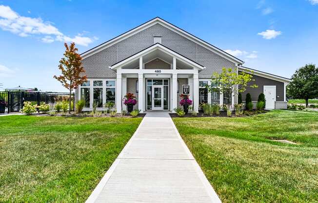 A building with a grey roof and a white door is surrounded by a grassy area and a concrete pathway leading to the entrance.