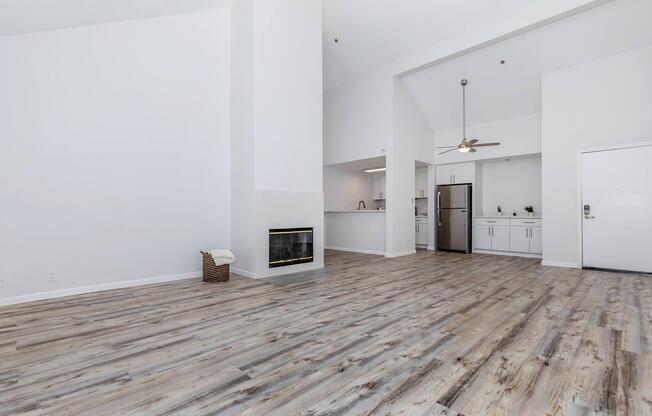 A modern, open-concept living space with light wood flooring, a white fireplace, and a ceiling fan. The kitchen area features stainless steel appliances and white cabinetry. The high ceiling and large windows create a bright atmosphere. A door leads to the entrance, offering a view of the living room.