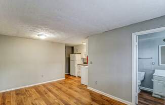 A room with wooden floors and a white ceiling with a kitchen area visible through the door.