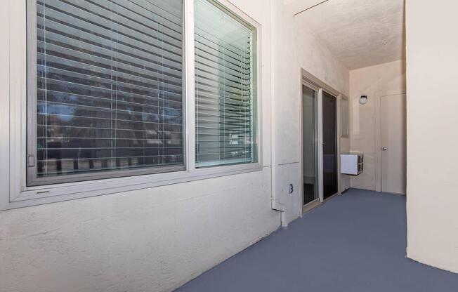 A view of a light-colored hallway with large windows featuring horizontal blinds. On the left, a window allows natural light to enter, while on the right, there are glass doors leading to an outdoor area. The floor is painted in a muted color, and the walls are smooth and textured.