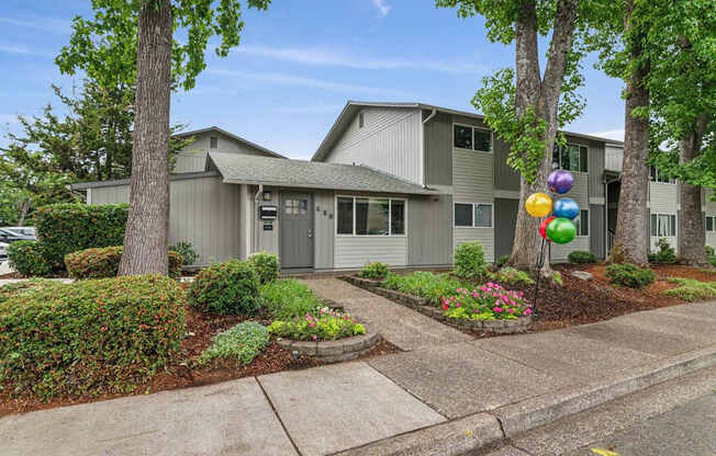 A house with a grey facade is surrounded by greenery and has a bunch of balloons tied to a tree.