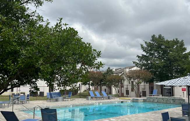 a swimming pool with blue chairs and trees and a cloudy sky