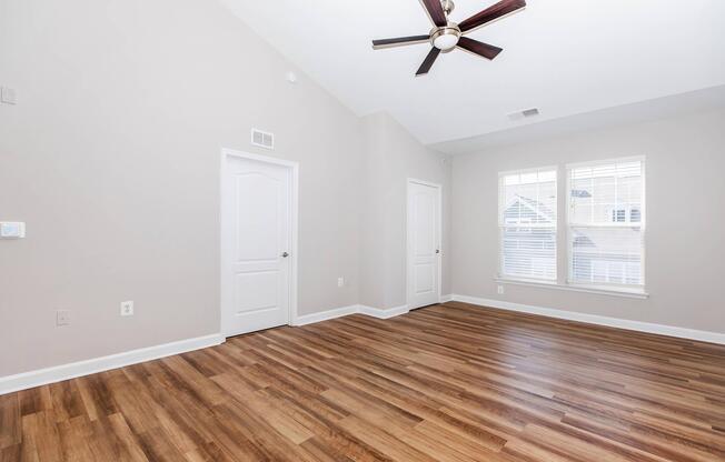Bright and spacious interior of a room with light gray walls and a vaulted ceiling, featuring a ceiling fan. The floor is covered with wooden laminate. Two doors are visible, one leading to a closet and another to an adjacent room. Large windows allow natural light, enhancing the open and airy atmosphere.