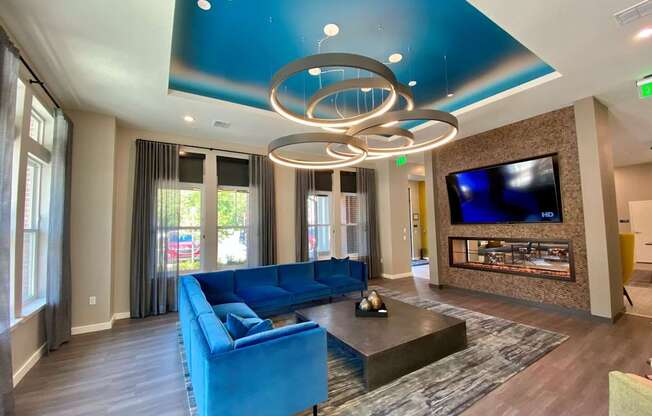 A clubhouse lobby with large windows, a comfortable L-shaped sofa, a large TV, and modern circular chandelier lighting.