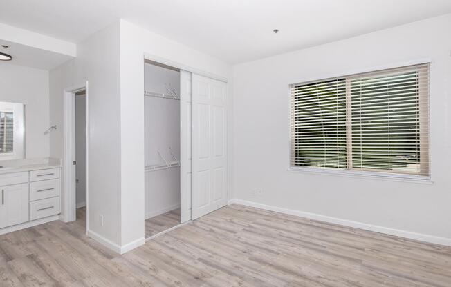 A bright, modern bedroom featuring light-colored walls and wooden flooring. There is a window with blinds allowing natural light, a small closet with sliding doors, and a sink area visible in the background. The space is uncluttered and designed for optimal use.