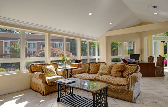 A sunlit resident lounge here at Adagio featuring large windows that fill the space with natural light, comfortable upholstered sofas and armchairs arranged around a decorative coffee table, and tile flooring throughout, while an adjacent desk area provides workspace and views of landscaped community buildings enhance the inviting, relaxed atmosphere.