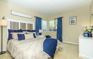 A cozy bedroom featuring a large bed with beige bedding and dark blue accents. The room has light-colored walls, two windows with blue curtains, and tasteful decor, including a bedside lamp and a dresser. A framed picture and decorative items add a personal touch to the space.