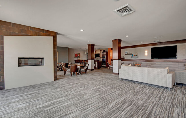 A spacious resident clubhouse here at Vue at Pinnacle Park with open lounge seating, a modern fireplace wall, dining tables, a large TV area, and a kitchen bar space for comfortable community gatherings.