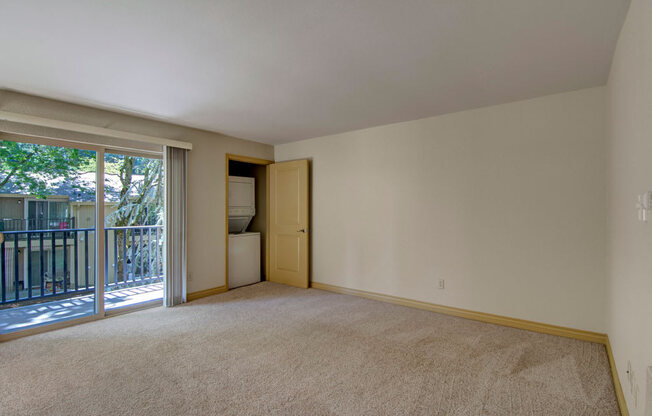 A spacious living room here at Asbury Park featuring plush carpet flooring, soft neutral walls, and a large sliding glass door that opens to a private balcony overlooking mature trees, along with an in-home laundry closet tucked behind a partially open door and an open layout offering ample wall space for seating and entertainment furniture.