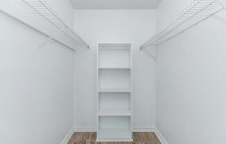 A clean, empty closet with white walls and wooden flooring. It features two wire shelves on either side and a vertical white storage unit in the center. The space is bright and uncluttered, ideal for organizing clothes or other items.