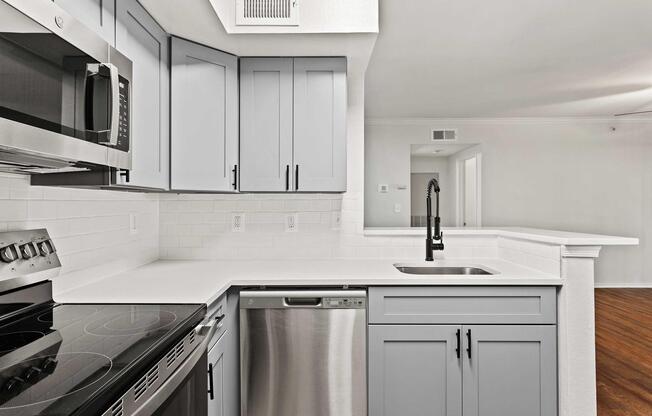 Modern kitchen featuring light gray cabinetry, a stainless steel microwave, oven, and dishwasher, with a sleek white countertop and a black faucet. The open layout connects to a brightly lit living area with hardwood flooring and neutral-colored walls.