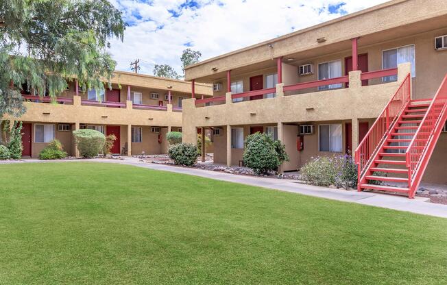 A view of a tidy apartment complex featuring two-story buildings with balconies. The buildings have a beige exterior and red accents, surrounded by well-maintained green lawns and shrubs. A staircase leads to the second floor, and the sky is partly cloudy.