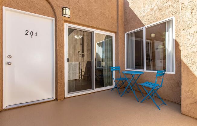 A cozy outdoor space featuring a light brown stucco wall with a white door labeled "203." There is a sliding glass door leading into a room, alongside two bright blue folding chairs and a small table, creating an inviting area for relaxation.