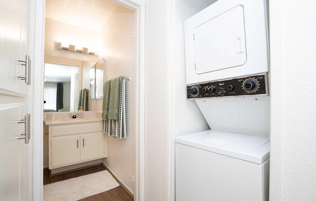 A view of a laundry area with a stacked washer and dryer. The space features a doorway leading to a well-lit bathroom, which includes a mirror, sink, and towel rack with green towels. The flooring is dark wood, and the walls are painted in light colors.