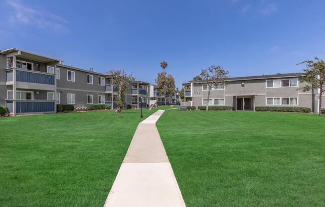 A well-kept residential complex featuring two-story apartment buildings arranged around a green lawn. A concrete pathway runs through the center, leading to the buildings. The sky is clear with a few clouds, and there are palm trees and landscaping around the area, creating a peaceful suburban environment.