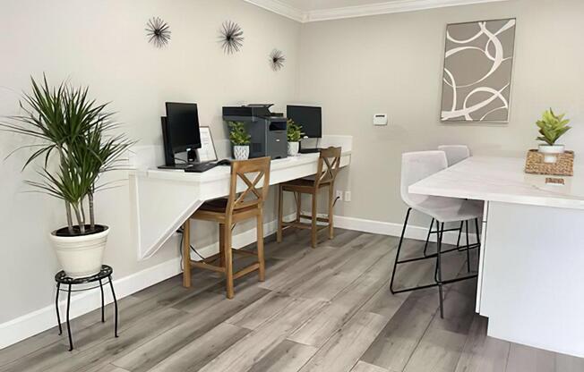 A modern workspace featuring two desks with computers and chairs, a printer, and decorative plants. Light-colored walls and wooden flooring create a bright and airy atmosphere. A large artwork piece hangs on the wall, adding a contemporary touch.