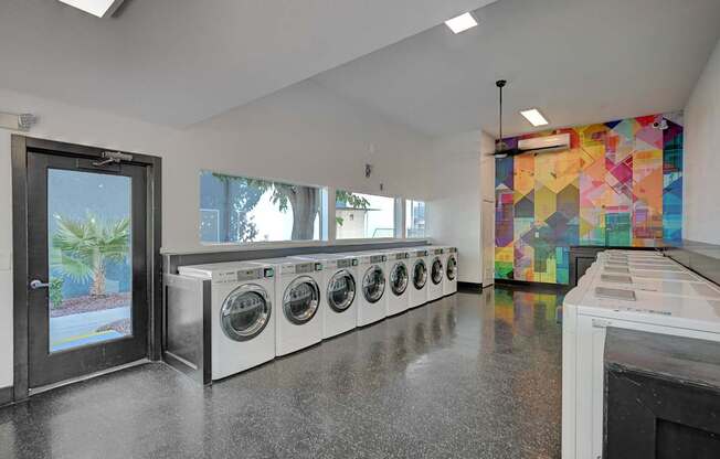 A laundry room with a row of washing machines and a colorful mosaic wall.