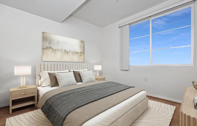 A modern bedroom featuring a bed with light-colored bedding and decorative pillows. There are two bedside tables with lamps, and a large window with a clear view of the sky. The walls are painted white, and there is an abstract artwork hanging above the bed.
