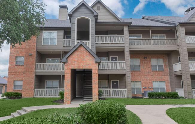A multi-story residential building featuring brick and siding exteriors. The entrance has a covered archway leading to a staircase. Balconies are visible on the front and sides of the building, surrounded by well-maintained green grass and landscaping under a partly cloudy sky.
