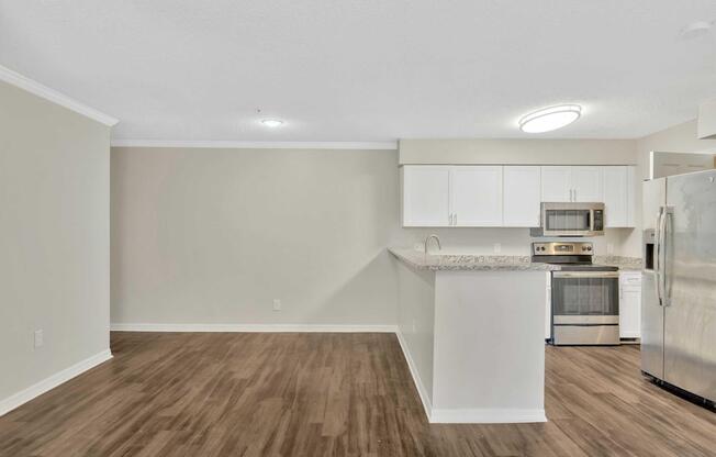 Modern kitchen and living area featuring light-colored walls, laminate flooring, and an open layout. The kitchen has white cabinets, stainless steel appliances, and a spacious countertop. A brightness from ceiling lights enhances the clean and inviting atmosphere.